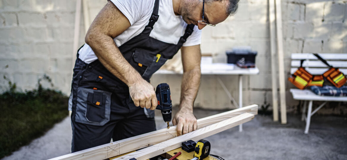 Mature male carpenter using electric drill on a wooden plank out